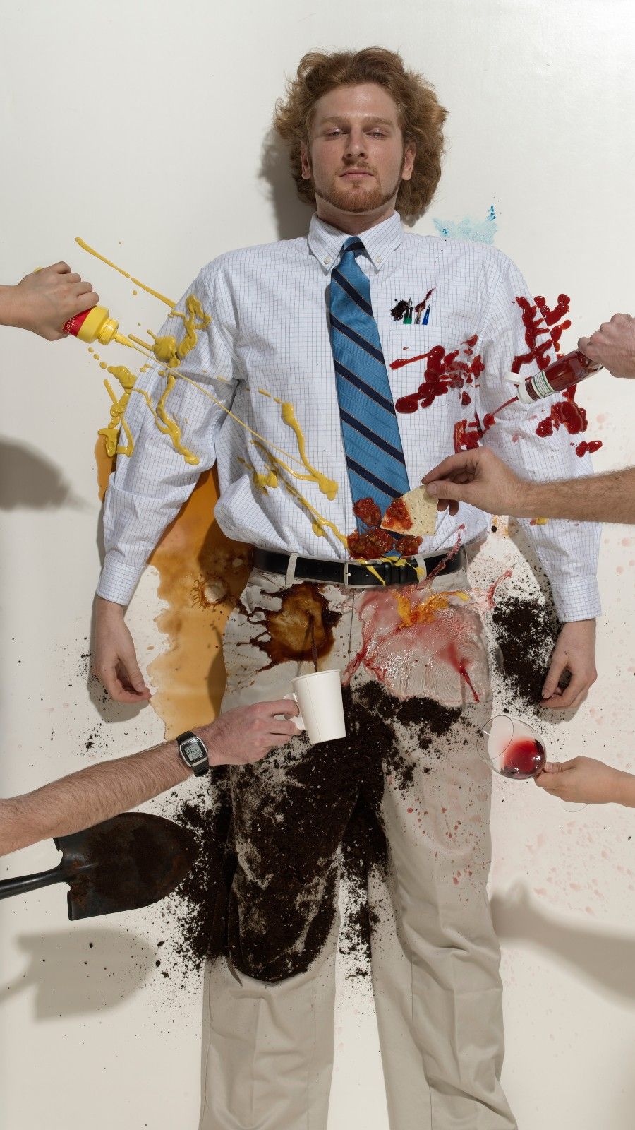 Homem de 20 anos deitado no chão vestindo uma camisa de botão, gravata e cáqui com pessoas servindo ketchup, mostarda, molho, café, sujeira e vinho em um fundo branco