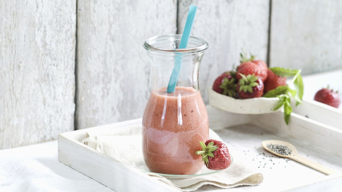 jahodové smoothie v pohári s modrou slamkou. Pohár je na bielom podnose vedľa misky jahôd